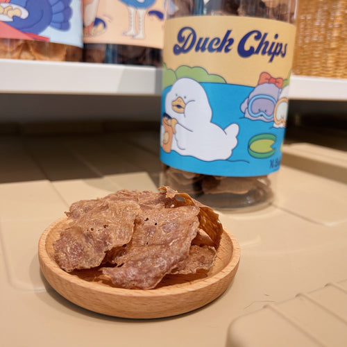 K Bakery Duck Chips — thin light-brown dried duck meat chips in a wooden dish, with the blue-labelled glass jar featuring a cartoon duck in the background